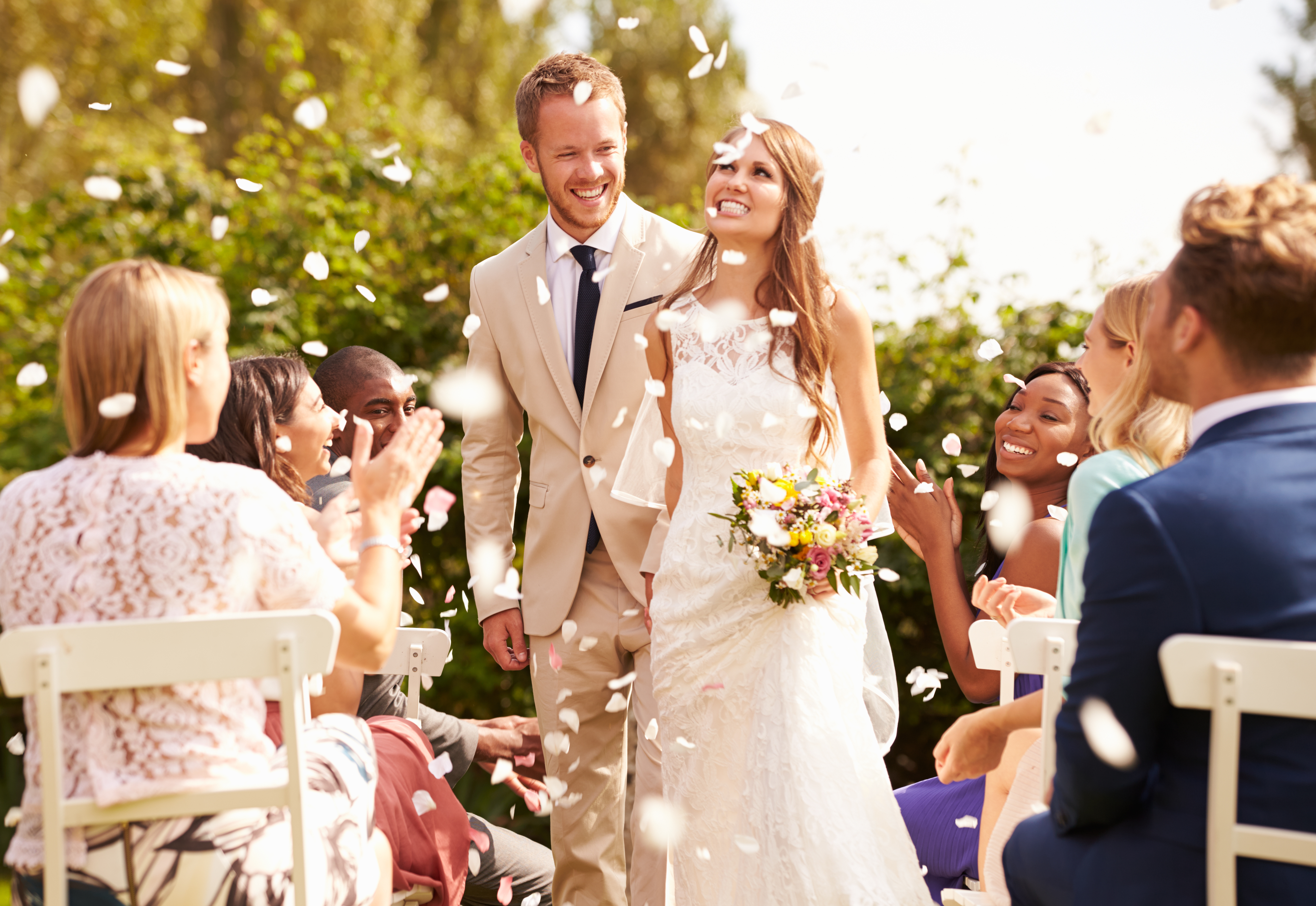 A beaming bride and groom walk hand-in-hand down the aisle, surrounded by joyful guests showered by tissue rose petals into the air, capturing the romance and celebration of a perfect wedding day.