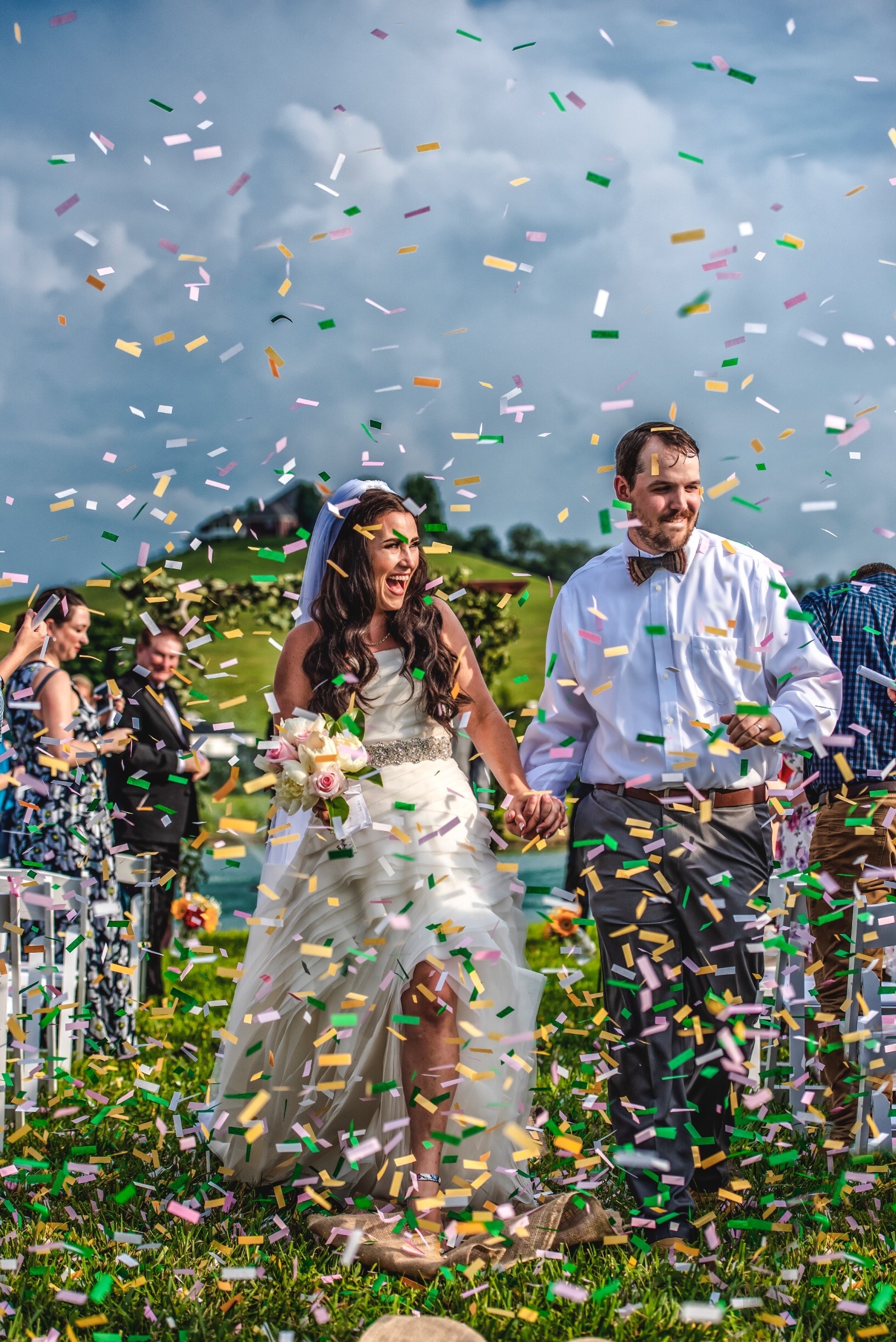 A joyful bride and groom walk down the aisle surrounded by vibrant, biodegradable confetti in the breeze, capturing the perfect outdoor wedding moment.